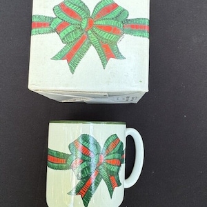 May include: A white ceramic mug with a green and red bow design. The mug has a green rim and handle.