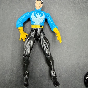 May include: A plastic action figure of a male superhero. The figure has a blue top with a white emblem, black pants, yellow gloves, and a yellow belt. The figure is standing with its legs apart.