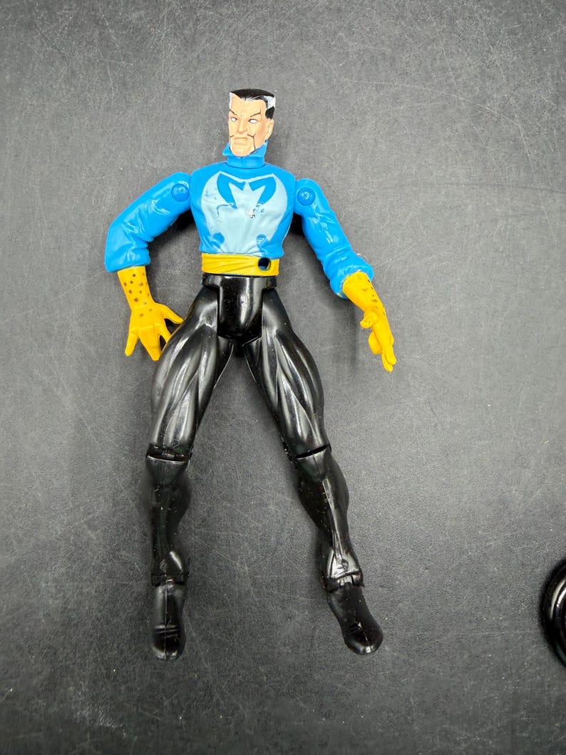 May include: A plastic action figure of a male superhero. The figure has a blue top with a white emblem, black pants, yellow gloves, and a yellow belt. The figure is standing with its legs apart.