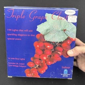May include: A box for a set of 144 indoor string lights shaped like grapes. The box is blue with a picture of a bunch of red grapes on a vine. The text on the box reads "Triple Grape Cluster", "144 Lights that will add sparkling elegance to that special event", "Lit with Rice Lights", "Includes UL Listed Low Voltage Adaptor", "Indoor", and "Green 21. PH.NF 727007 £33.22".