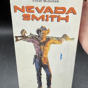 May include: A vintage paperback book titled "NEVADA SMITH" by Steve McQueen. The cover features a shirtless man in a cowboy hat holding a rifle. The title is in orange block letters.