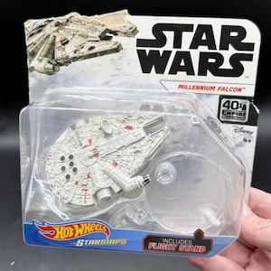Könnte beinhalten: Ein Star Wars Millennium Falcon Hot Wheels Starships Spielzeug in Originalverpackung. Das weiß-graue Raumschiff hat rote Markierungen und enthält einen durchsichtigen Flugständer aus Kunststoff. Die Karte zeigt das Star Wars Logo und Text.