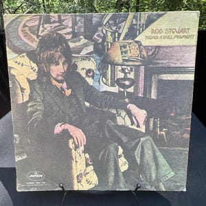 May include: Vintage vinyl record album cover featuring Rod Stewart. The album cover has a faded, vintage aesthetic. The text "Rod Stewart Never a Dull Moment" is visible. The album cover is displayed on a black metal stand.