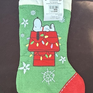 May include: Green Christmas stocking featuring Snoopy on his doghouse decorated with Christmas lights. The stocking has a red toe and a white cuff with the words "A HOL" and snowflakes. A tag is attached to the top.