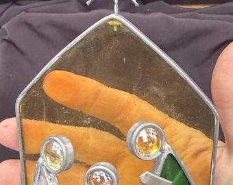 Puede incluir: Un adorno de natividad de vidrio tintado con tres figuras de cabezas circulares y cuerpos geométricos en blanco, naranja y verde. La pieza tiene forma de casa con un fondo amarillo claro y una estrella de alambre trenzado en la parte superior.