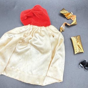 May include: A doll's costume set featuring a red velvet hood, a cream-colored satin dress, a gold-colored belt, a gold-colored cuff, and a pair of black shoes. The costume is likely vintage and appears to be in good condition.
