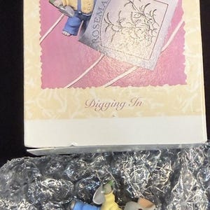 Può includere: Un ornamento Hallmark Keepsake con un topo in tuta blu e camicia gialla, che tiene in mano un cartello con la scritta "Rosemary". L'ornamento è confezionato in una scatola decorativa con la scritta "Digging In".