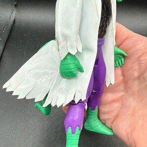 K&ouml;nnte beinhalten: Eine gr&uuml;ne, echsenartige Actionfigur mit einem wei&szlig;en Laborkittel und einer lila Hose. Die Figur hat einen gr&uuml;nen Kopf mit roten Augen und einem weit ge&ouml;ffneten Mund. Die H&auml;nde und F&uuml;&szlig;e der Figur sind gr&uuml;n.