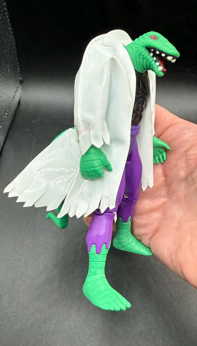 K&ouml;nnte beinhalten: Eine gr&uuml;ne, echsenartige Actionfigur mit einem wei&szlig;en Laborkittel und einer lila Hose. Die Figur hat einen gr&uuml;nen Kopf mit roten Augen und einem weit ge&ouml;ffneten Mund. Die H&auml;nde und F&uuml;&szlig;e der Figur sind gr&uuml;n.