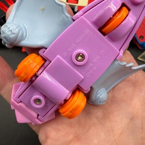May include: A purple and light blue toy vehicle with orange wheels. The toy has the text "&copy; 1995 Marvel" and "China WH 25 Chine" printed on it. The toy is held in a hand.