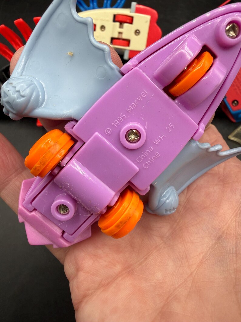 May include: A purple and light blue toy vehicle with orange wheels. The toy has the text "&copy; 1995 Marvel" and "China WH 25 Chine" printed on it. The toy is held in a hand.
