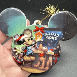 Puede incluir: Adorno navideño con forma de Mickey Mouse que representa a Mickey y Pluto en una tienda. Mickey lleva un gorro de Papá Noel y un peto azul. El adorno tiene una cuerda dorada para colgar y se ven las palabras "CLOCK SHOP".