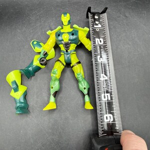 May include: A yellow and green action figure, approximately 5 inches tall, with articulated limbs and a weapon attachment. The figure is positioned next to a measuring tape.