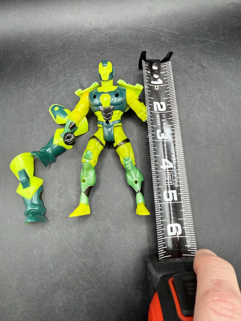 May include: A yellow and green action figure, approximately 5 inches tall, with articulated limbs and a weapon attachment. The figure is positioned next to a measuring tape.
