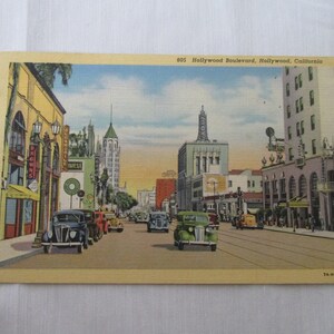 May include: Vintage postcard depicting Hollywood Boulevard in Hollywood, California. The image shows classic cars on the street, buildings with vintage signage, and a clear blue sky. The postcard has a yellowed, aged appearance.