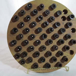 May include: A round wooden display board with numerous dark-colored bottle stoppers. The board has handwritten labels and several empty holes. The board is supported by a gold-colored wire stand.