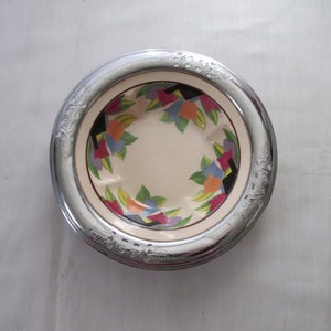May include: A decorative silver-toned ashtray with a floral design. The inner bowl features a cream-colored background with a colorful floral pattern in shades of blue, orange, purple, and green. The rim has an embossed floral pattern.