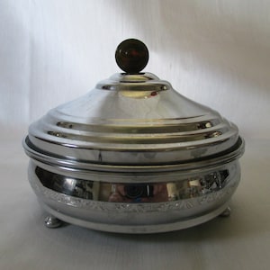 May include: A silver-colored, lidded serving dish with a decorative floral pattern around the base. The lid is topped with a small, round, dark-colored knob. The dish has three small feet.