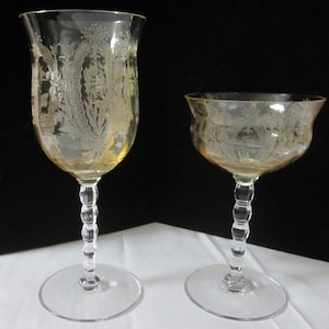 May include: Two etched glass goblets with a yellow tint. The taller goblet has a tulip shape, while the shorter one is a champagne coupe. Both feature intricate floral and landscape designs, with clear stems and bases.