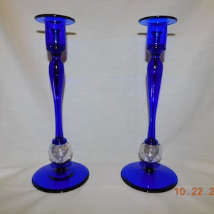May include: Two cobalt blue glass candlesticks. Each candlestick has a round base, a slender stem, and a clear crystal ball accent. The top of each candlestick has a flat, circular platform to hold a candle.