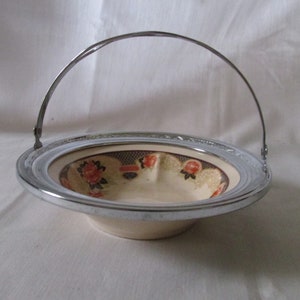 May include: A small, round serving dish with a silver-colored metal rim and handle. The interior features a floral pattern with red and orange flowers on a cream background. The dish is likely vintage.