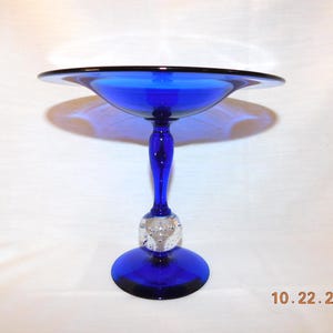 May include: A cobalt blue glass pedestal bowl with a wide, shallow dish and a rounded base. The stem features a clear, decorative ball. The bowl is likely used for serving fruit or displaying decorative items.