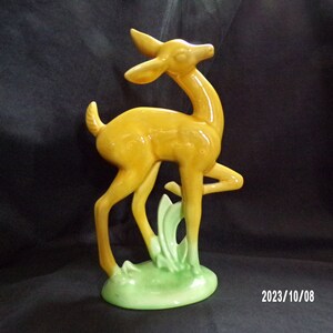 May include: A ceramic figurine of a deer in a standing pose. The deer is a glossy yellow color with a slender neck and legs. It stands on a small green base with stylized grass. The figurine is a decorative item.