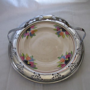 May include: A vintage serving dish with a cream-colored center featuring a floral design in shades of blue, orange, and purple. The dish is encircled by a silver-colored metal rim with decorative cutouts and handles.