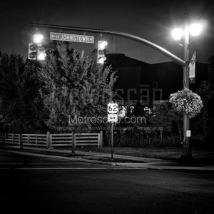 Puede incluir: Una foto en blanco y negro de una intersección de calles por la noche. Un letrero de calle dice "US-62 JOHNSTOWN RD". Un semáforo está en verde. Un letrero con una flecha blanca y el número 62 es visible. Una farola está encendida.