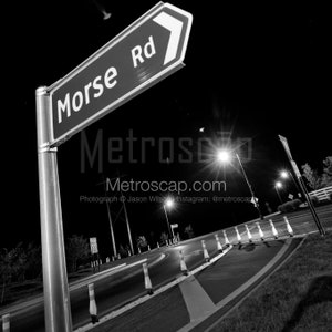 Puede incluir: Una fotografía en blanco y negro de una señal de calle que dice "Morse Rd" con una flecha apuntando hacia la derecha. La señal está montada en un poste de metal. La calle está vacía y hay farolas a ambos lados de la carretera.