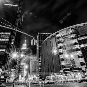 Puede incluir: Una fotografía en blanco y negro de una calle de la ciudad por la noche. El Empire State Building es visible en la distancia. El Madison Square Garden está en primer plano con una gran pancarta que dice "BE ANYONE, HAVE IT ALL".
