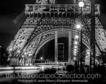 Puede incluir: Una fotografía en blanco y negro de la Torre Eiffel de noche. La torre está iluminada y hay gente caminando frente a ella. La foto fue tomada desde el nivel de la calle y muestra la torre de abajo hacia arriba.