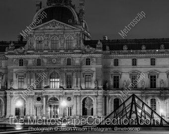 Puede incluir: Una fotografía en blanco y negro del Museo del Louvre en París, Francia, de noche. La icónica pirámide de vidrio está en primer plano, con la fachada del museo en el fondo.