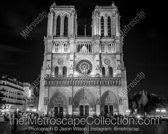 Puede incluir: Una fotografía en blanco y negro de la Catedral de Notre Dame en París, Francia, de noche. La catedral está iluminada con luces, y hay gente caminando frente a ella.