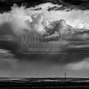 Peut inclure: Une photographie en noir et blanc d'une grande formation nuageuse sombre et orageuse au-dessus d'un champ plat et herbeux. Le ciel est rempli de nuages ​​dramatiques et tourbillonnants, et le sol est presque entièrement obscurci par les nuages ​​bas. L'image est prise de loin, et l'horizon est visible à l'arrière-plan.