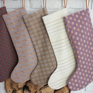 Peut inclure: Cinq chaussettes de Noël accrochées à un mur de briques blanches. Les chaussettes sont en tissu et présentent différents motifs, notamment des motifs géométriques, des rayures et du tartan. Les chaussettes sont toutes dans des tons de brun, beige et violet.