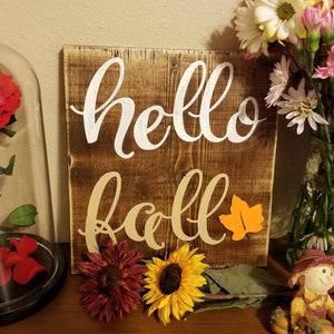 Può includere: Un cartello di legno con le parole "hello fall" scritte in vernice bianca e crema. Il cartello ha una finitura rustica marrone e un piccolo ritaglio di foglia arancione.