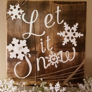 Può includere: Un cartello in legno rustico con le parole "Let it Snow" in lettere bianche. Il cartello è decorato con fiocchi di neve bianchi e spago.