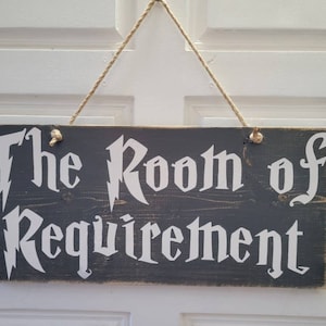 May include: A gray wooden sign with white lettering that reads "The Room of Requirement". The sign is hanging from a string.