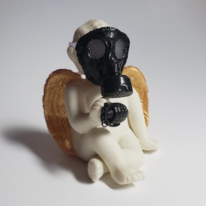 May include: A ceramic cherub figurine with golden wings, wearing a black gas mask and holding a black grenade. The cherub is sitting down with its legs crossed. The background is white.