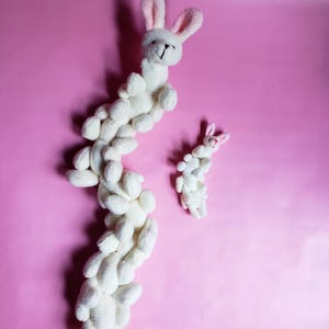 May include: Two white plush bunny toys on a pink background. The larger bunny is curled into a long, winding shape, while the smaller bunny is standing upright.
