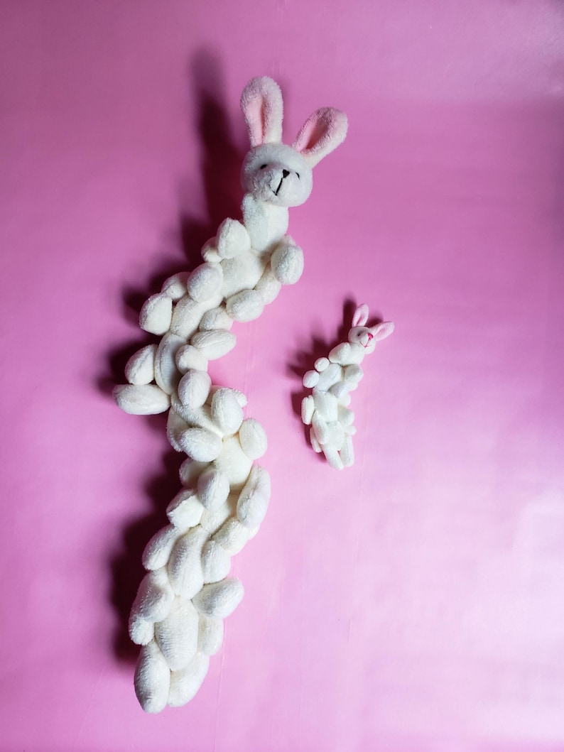 May include: Two white plush bunny toys on a pink background. The larger bunny is curled into a long, winding shape, while the smaller bunny is standing upright.