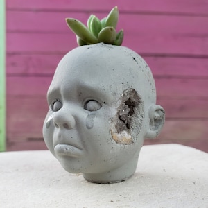 Puede incluir: Una maceta de hormigón gris con forma de cabeza de muñeca llorando, con una planta suculenta encima. La cabeza tiene una gran abertura en el lateral que revela cristales. La muñeca tiene una lágrima en la mejilla.