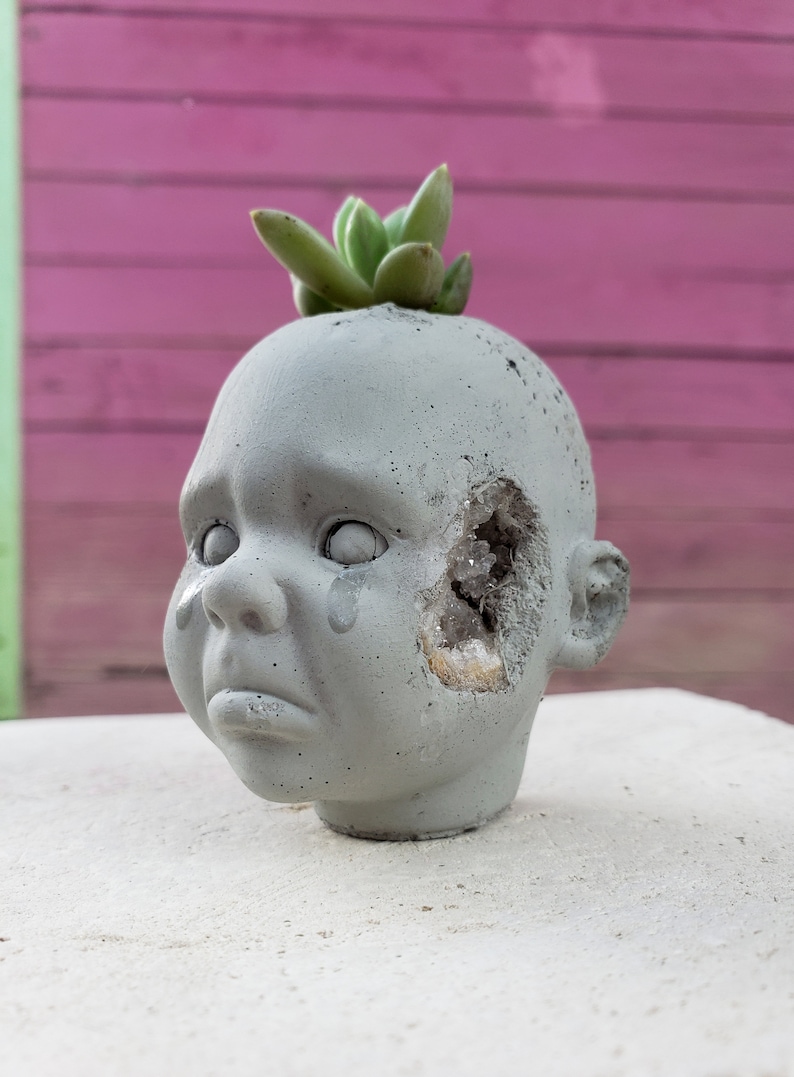 May include: A grey concrete planter in the shape of a crying baby doll's head, with a succulent plant on top. The head has a large opening on the side revealing crystals. The doll has a tear on its cheek.