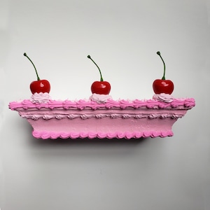Op de afbeelding: Een roze, rechthoekige sculptuur die op een cake lijkt, versierd met drie rode kersen. Elke kers zit bovenop een toefje witte glazuur. De sculptuur heeft een gelaagd ontwerp met een decoratieve, getextureerde rand.