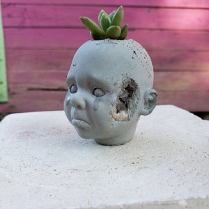 May include: A grey concrete planter in the shape of a baby's head, with a succulent plant growing from the top. The head has a distressed look with a hole showing a crystal-like interior. The planter is on a white surface.