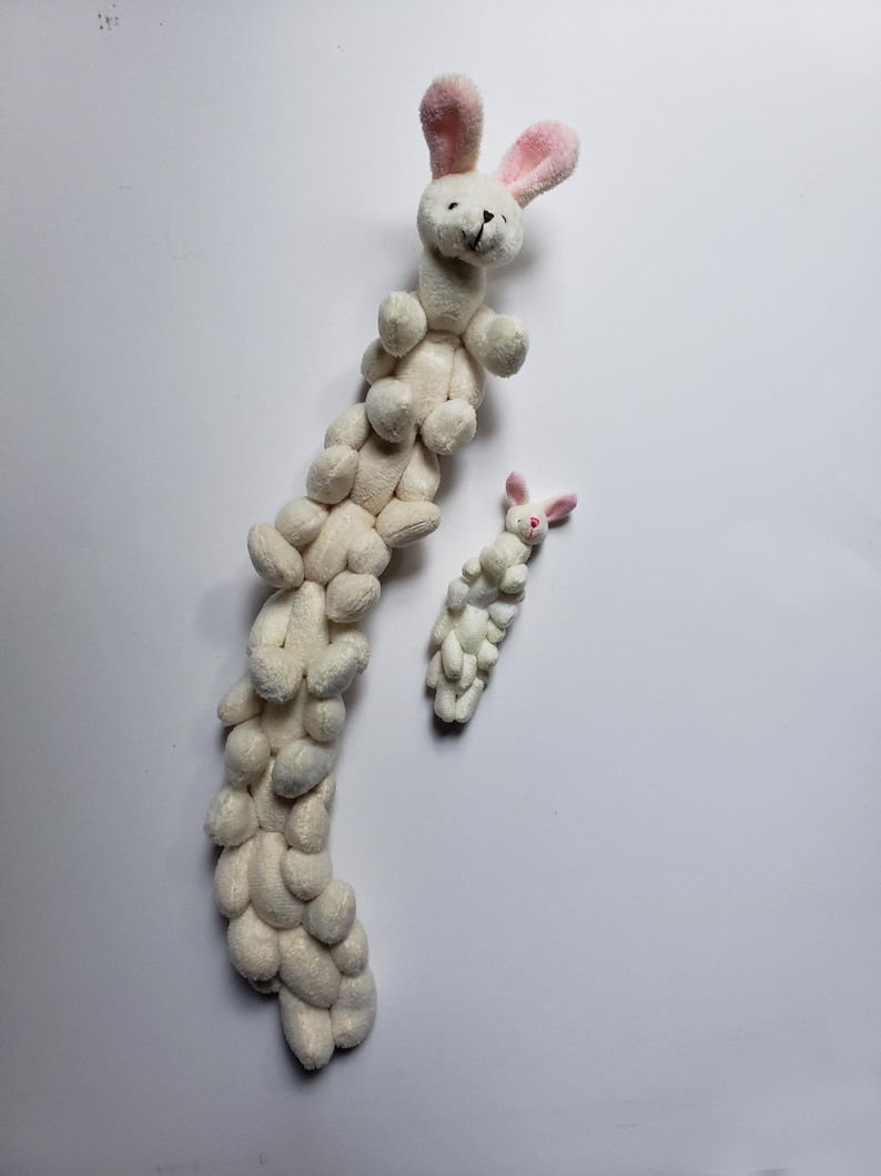 May include: A white plush bunny toy with pink ears is stacked on top of other white plush bunny toys, forming a long chain. A smaller white plush bunny toy with pink ears is also pictured.