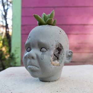 May include: A grey concrete baby head planter with a hole in the side and a succulent plant on top. The head has a sad expression with tears. The planter is on a white surface.