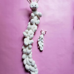 May include: A white plush bunny toy with a long, segmented body made of smaller bunny shapes. The toy is on a pink background.