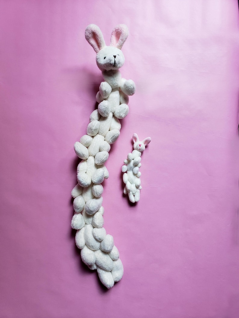 May include: A white plush bunny toy with a long, segmented body made of smaller bunny shapes. The toy is on a pink background.